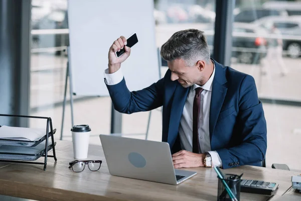 Messa a fuoco selettiva di uomo d'affari sconvolto in tuta che tiene smartphone vicino al computer portatile e alla tazza di carta in ufficio — Foto stock