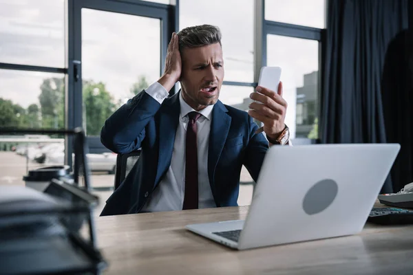 Selective focus of surprised businessman using smartphone near laptop in office — Stock Photo