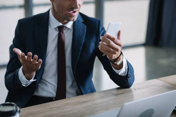 Vue recadrée de l'homme d'affaires surpris à l'aide d'un smartphone et geste près d'un ordinateur portable — Photo de stock