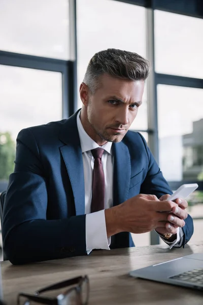 Enfoque selectivo de hombre de negocios guapo sosteniendo el teléfono inteligente y mirando a la cámara - foto de stock