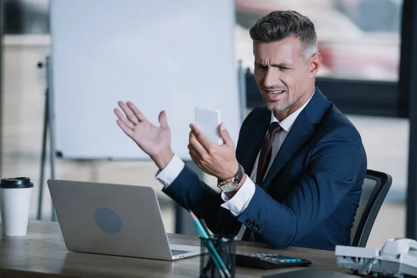 Enfoque selectivo del hombre molesto gesto cerca de la computadora portátil mientras sostiene el teléfono inteligente - foto de stock