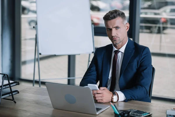 Selektiver Fokus eines gutaussehenden Mannes, der in die Kamera schaut, während er sein Smartphone in der Nähe des Laptops hält — Stockfoto