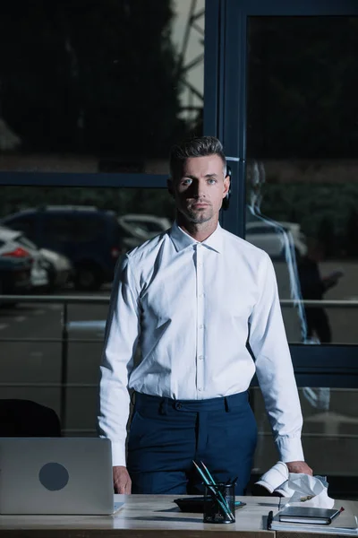 Guapo hombre de negocios mirando a la cámara mientras está de pie cerca del escritorio - foto de stock