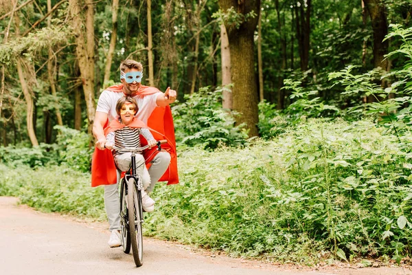 Padre y niño montar en bicicleta alrededor del bosque en trajes de superhéroe, mientras que papá señala con el dedo - foto de stock