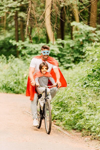 Повнометражний вид на батька і дитину, їзда на велосипеді навколо лісу в костюмах супергероїв — стокове фото