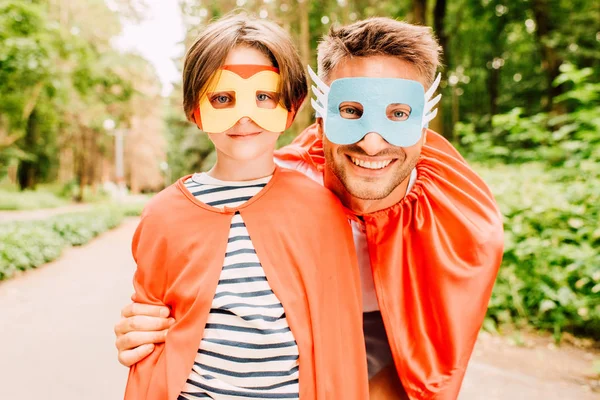 Pai feliz e filho de pé em capas vermelhas e máscaras de super-herói, sorrindo e olhando para a câmera — Fotografia de Stock