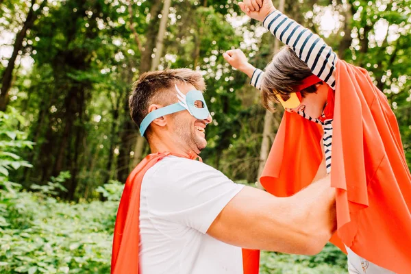 Vue latérale du père en costume de super-héros tenant le fils dans les mains pendant que l'enfant rit et regarde papa — Photo de stock