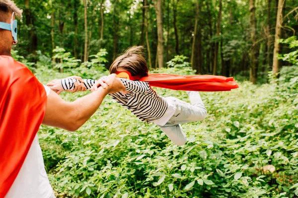 Padre en traje de superhéroe girando alrededor de hijo en capa roja - foto de stock