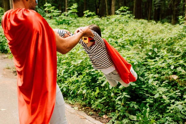 Father in red superhero cloak spinning around little son — Stock Photo