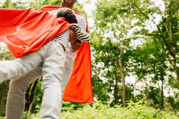 Baixo ângulo de visão do pai girando menino em manto de super-herói vermelho — Fotografia de Stock