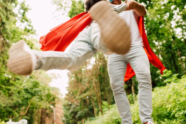 Baixo ângulo de visão do pai sorrindo enquanto girando menino em manto de super-herói vermelho — Fotografia de Stock