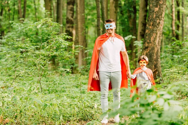 Pleine longueur vue de père et fils en costumes de super-héros tenant la main tout en se tenant dans la forêt verte — Photo de stock