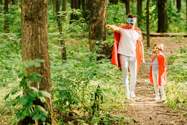 Ganzkörperansicht von Vater und Sohn, die in Superheldenkostümen im Wald stehen, während Papa mit dem Finger zeigt — Stockfoto