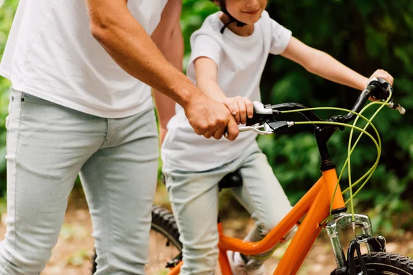 Vista recortada de hijo montar en bicicleta y padre sosteniendo asas para ayudar a niño a montar - foto de stock
