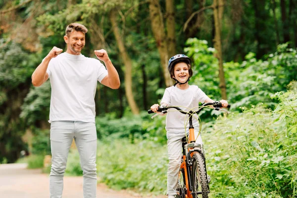 Vater lächelt und jubelt Sohn zu, während Junge Fahrrad fährt und in die Kamera schaut — Stockfoto