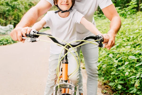 Vista ritagliata del padre che tiene in mano le maniglie della bicicletta mentre è seduto sorridente e seduto con casco in bicicletta — Foto stock
