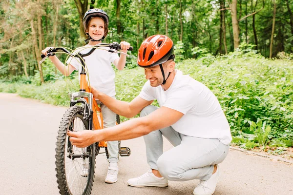 Отец в шлеме проверяет колесо велосипеда, пока сын стоит в шлеме и смотрит в сторону — стоковое фото