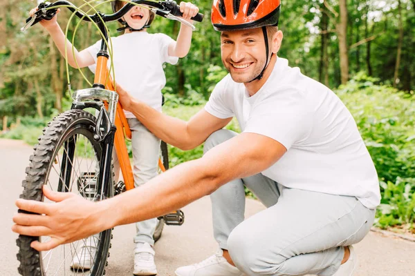 Vista recortada del hijo de pie y sosteniendo asas de bicicleta mientras el padre revisa la rueda - foto de stock
