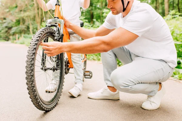 Vista cortada do pai no capacete verificando roda de bicicleta, enquanto o filho de pé acima da bicicleta — Fotografia de Stock