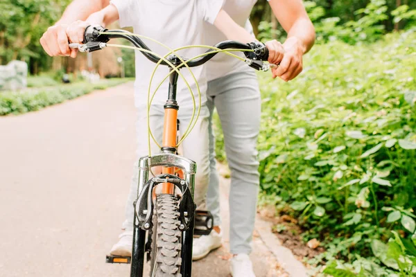 Cropped view of boy sitting on bicycle and father holding handles — Stock Photo