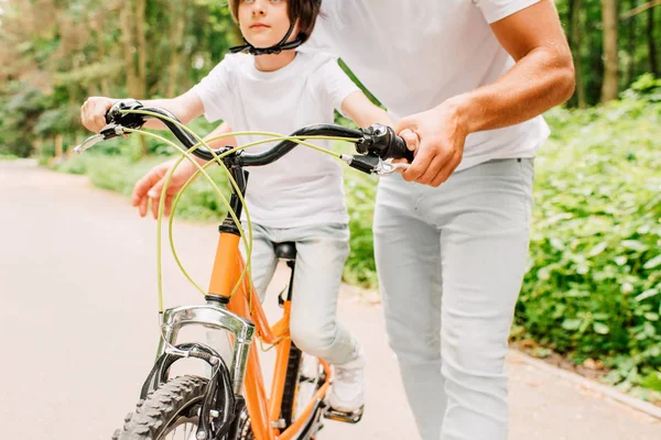 Vue recadrée du garçon dans le casque assis sur le vélo et le père tenant la poignée avec la main — Photo de stock