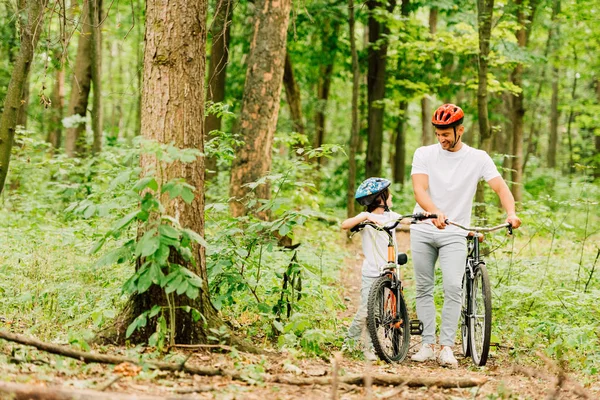 Повнометражний вид батька і сина в шоломах, що йдуть з велосипедами в лісі — стокове фото
