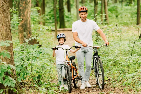 Ganzkörperansicht von Vater und Sohn, die mit Fahrrädern im Wald spazieren gehen und in die Kamera schauen — Stockfoto