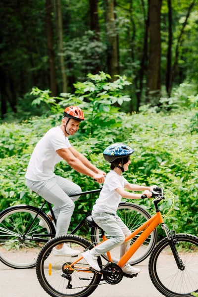 Вид отца и сына в полный рост, катающихся на велосипедах по дороге возле леса, пока отец смотрит на ребенка — стоковое фото
