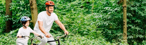 Prise de vue panoramique du père et du fils souriant en vélo dans la forêt — Photo de stock