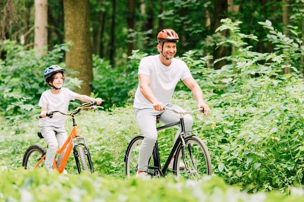 Foco seletivo de pai e filho sorrindo e olhando para a frente ao andar de bicicletas na floresta — Fotografia de Stock