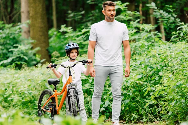 Vista completa del padre y el hijo de pie en la carretera cerca del bosque y mirando hacia adelante mientras el hijo sostiene la bicicleta - foto de stock