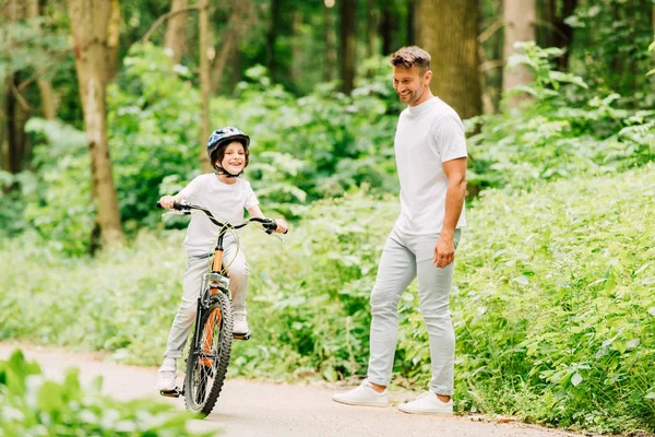 Visão de comprimento total da criança andando de bicicleta, enquanto o pai de pé perto e olhando para o filho — Fotografia de Stock