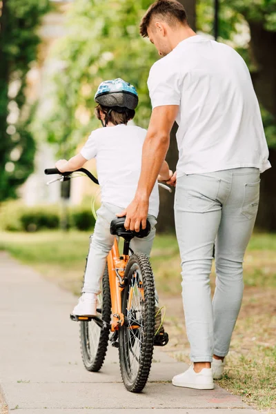 Ganzkörperansicht des Vaters, der dem Sohn hilft, mit dem Fahrrad zu fahren, indem er den Sitz des Fahrrads hält — Stockfoto