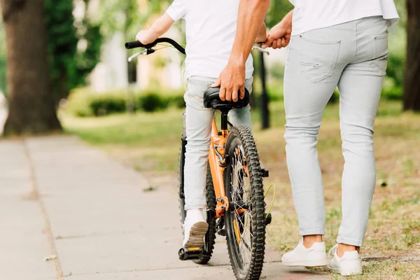 Vue recadrée du père aidant son fils à monter à vélo en tenant assis de vélo — Photo de stock