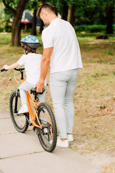 Vue arrière du père aidant son fils à monter à vélo en tenant assis de vélo — Photo de stock