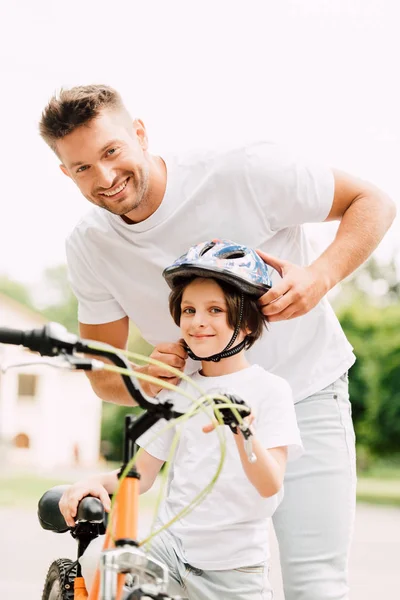 Selektiver Fokus von Vater und Sohn, die in die Kamera schauen, während Papa dem Jungen einen Helm aufsetzt — Stockfoto