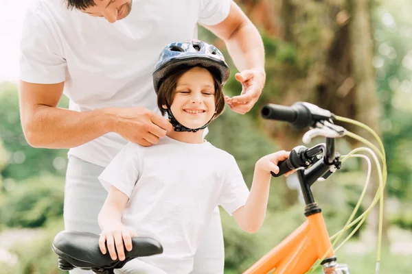 Vue recadrée du père portant un casque sur son fils tandis que le garçon souriant et debout près du vélo — Photo de stock