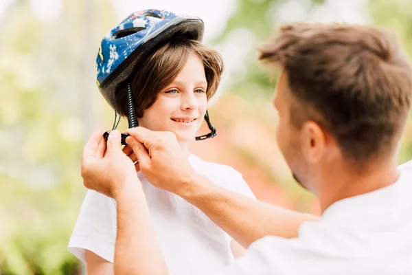 Избирательный фокус счастливого сына, смотрящего на отца в то время как отец надевает шлем — стоковое фото