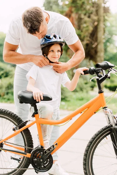 Ganzkörperansicht des Vaters, der dem Sohn einen Helm aufsetzt, während der Junge neben dem Fahrrad steht und wegschaut — Stockfoto