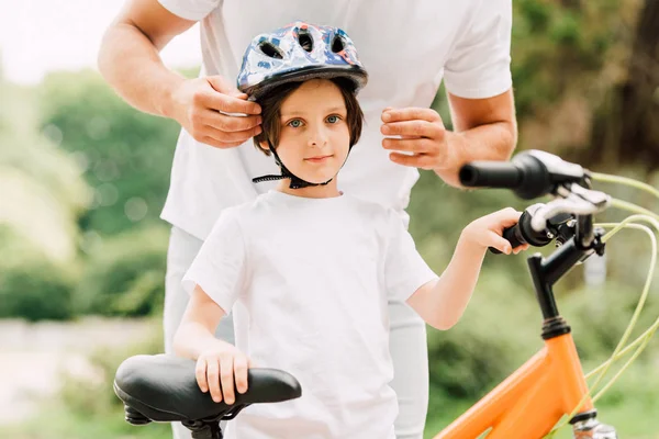 Ausgeschnittene Ansicht eines Jungen, der in die Kamera schaut, während der Vater seinem Sohn einen Helm aufsetzt — Stockfoto