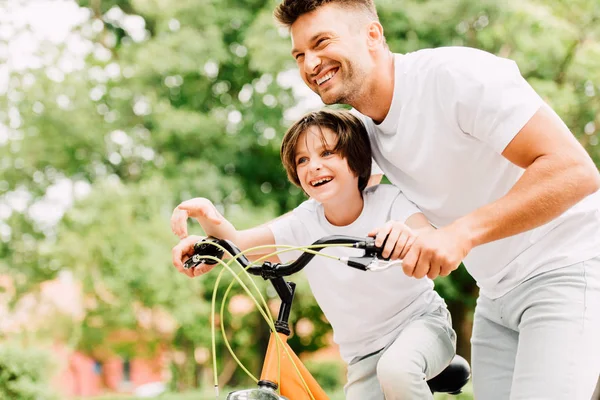 Feliz padre e hijo mirando hacia adelante mientras niño señalando con el dedo y papá ayudar a niño a montar en bicicleta - foto de stock