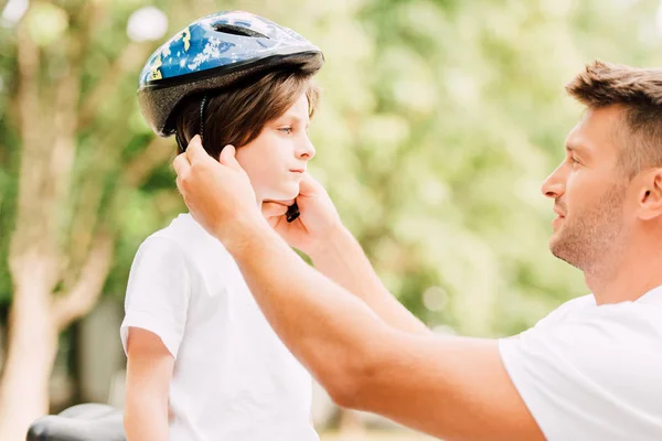 Side view of father putting helmet on son while boy looking at dad — Stock Photo