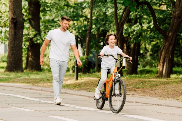 Pleine longueur vue de fils équitation vélo tandis que père marche après enfant — Photo de stock