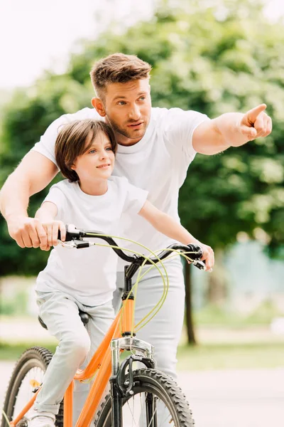 Padre e hijo mirando hacia adelante mientras el niño montar en bicicleta y papá señalando con el dedo - foto de stock