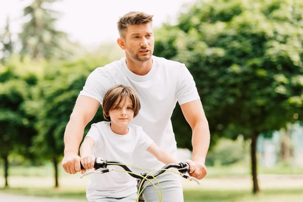 Father looking forward while son sitting on bicycle and looking at camera — Stock Photo