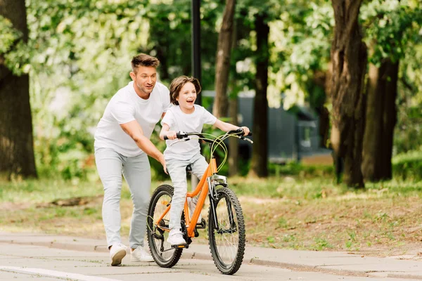 Longitud completa vista de padre empujando bicicleta mientras hijo deshaciéndose en bicicleta - foto de stock