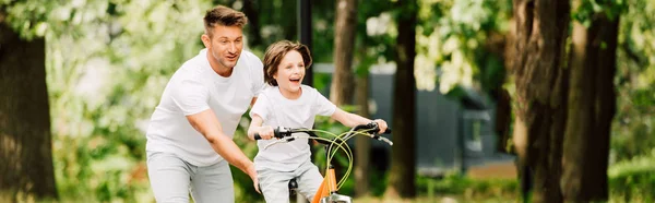 Plan panoramique de père poussant vélo tandis que son fils vélo — Photo de stock