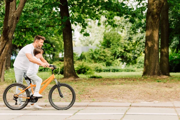 Vista lateral del padre ayudando a su hijo a montar en bicicleta mientras niño sentado en bicicleta - foto de stock