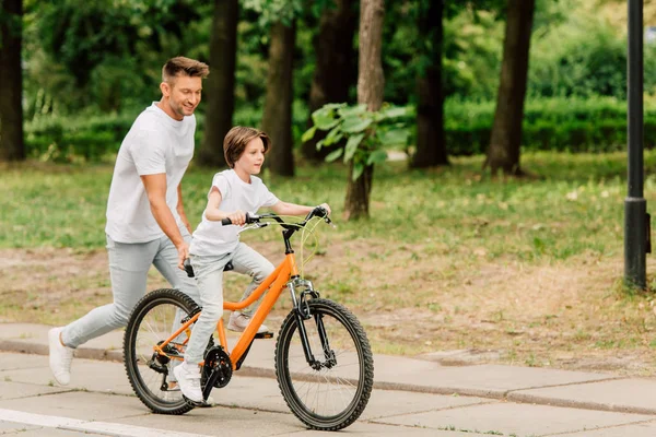Full length view of father pushing sit of bike and running after son while kid riding bicycle — Stock Photo