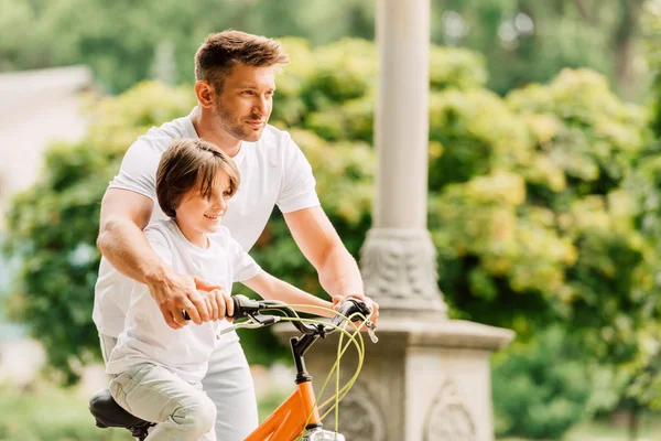 Enfoque selectivo de padre e hijo mirando hacia adelante mientras el niño montar en bicicleta y papá ayudar a niño - foto de stock